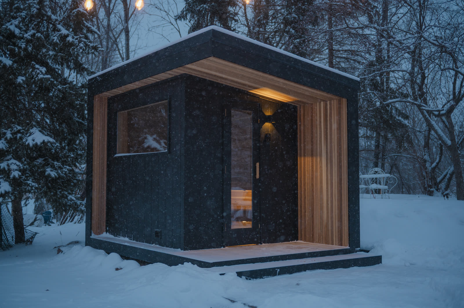 The black sauna at dusk under string lights, snow on the ground