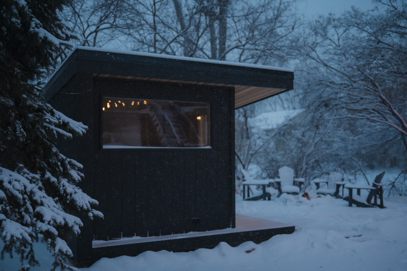 The side of the sauna with its window lit, chairs in the snow