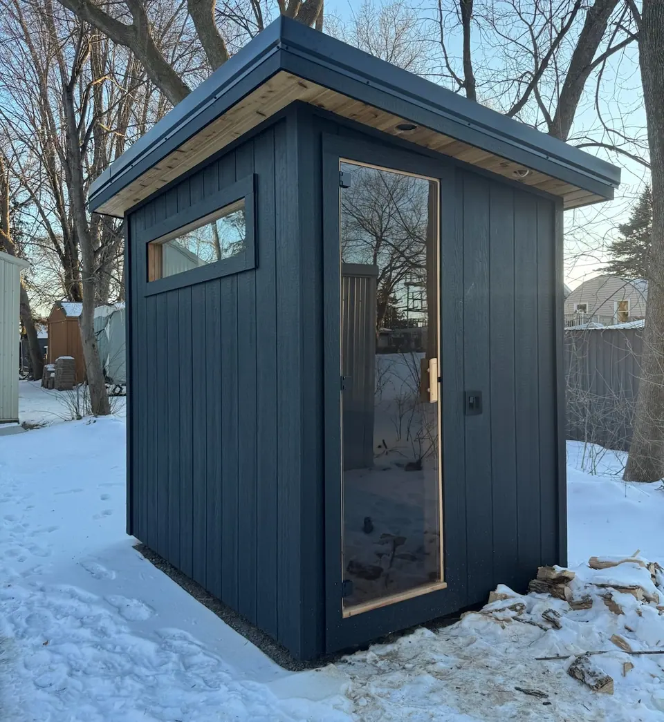 Black Kota sauna in the snow with split firewood stacked beside it