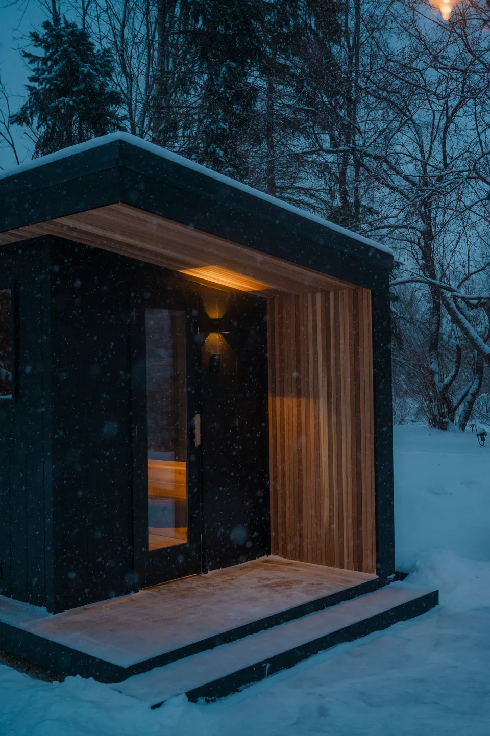 Cedar-lined entry of the black Tuli sauna, sconce lit at dusk in falling snow