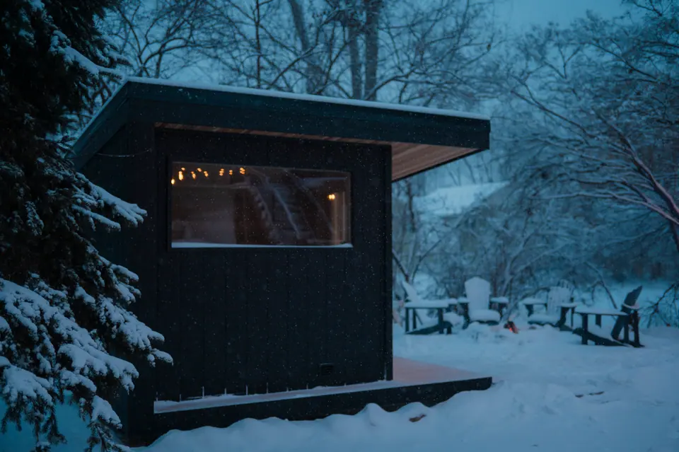 Side of the Tuli sauna at dusk with its window lit and chairs in the snow