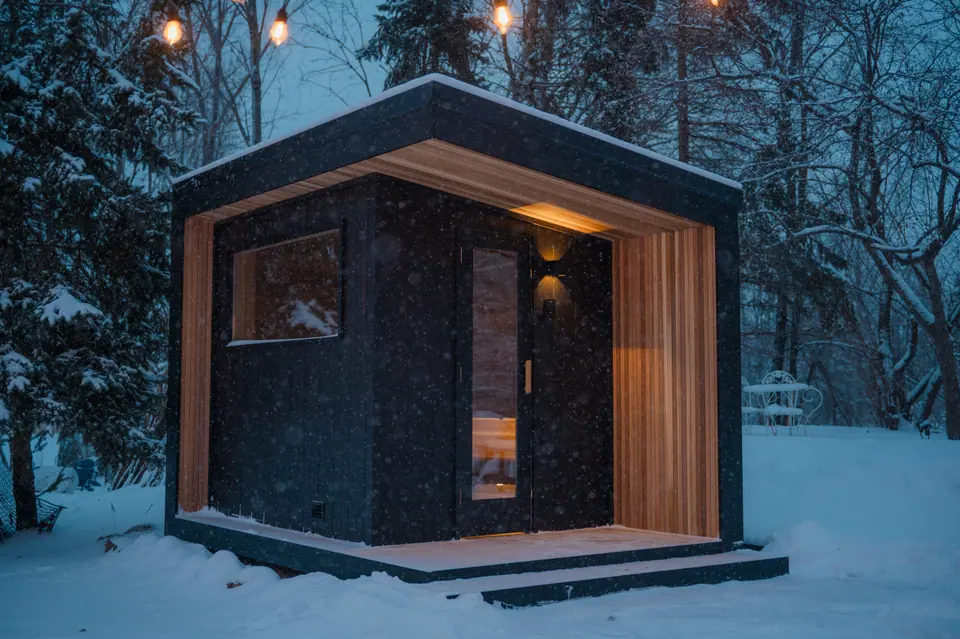 Black Tuli sauna with a cedar entry at dusk, snow on the ground and string lights overhead